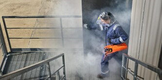 Person wearing protective gear and a respirator carrying an orange safety device while moving up metal stairs through a smoky industrial environment.

