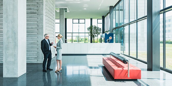 Wide shot of modern office building lobby surrounded by walls of windows with two businesspeople conversing
